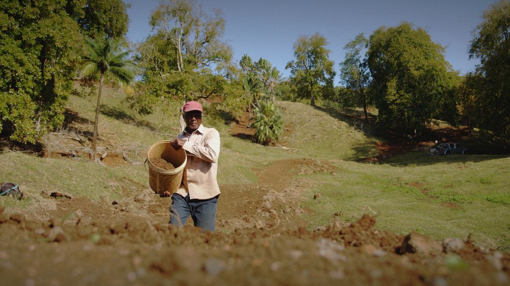 Patrick carries a basket of earth across the hillside at Mo’Bike, trees and the trail behind him