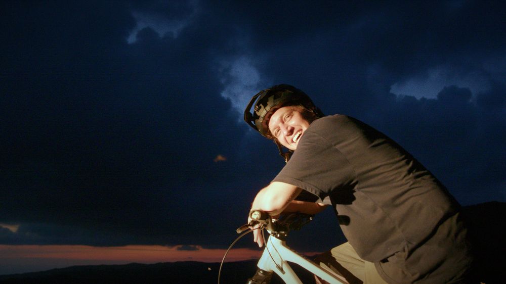 Henri on the bike at dusk, smiling, storm clouds above
