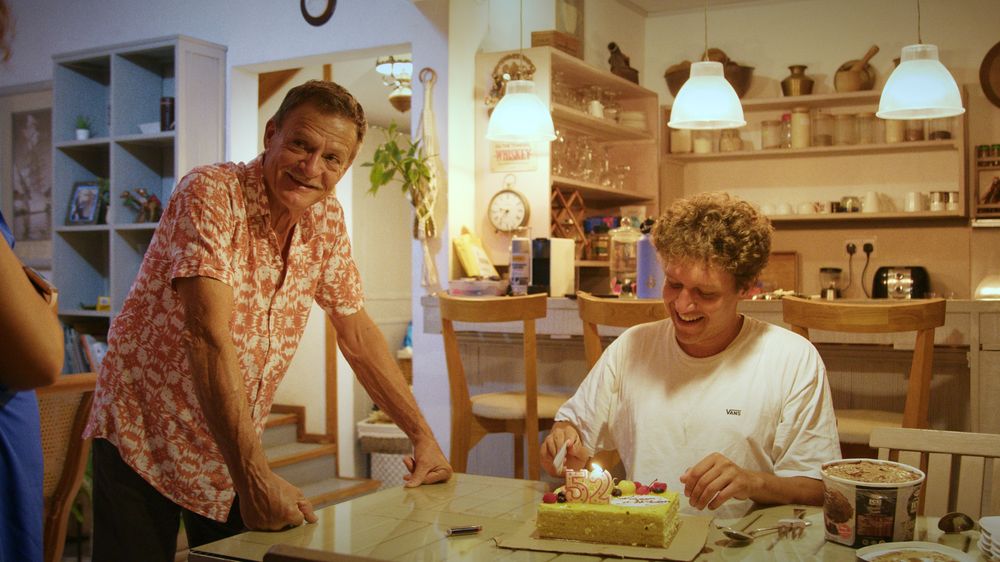 Henri laughs at a kitchen table over a birthday cake while an older man looks on