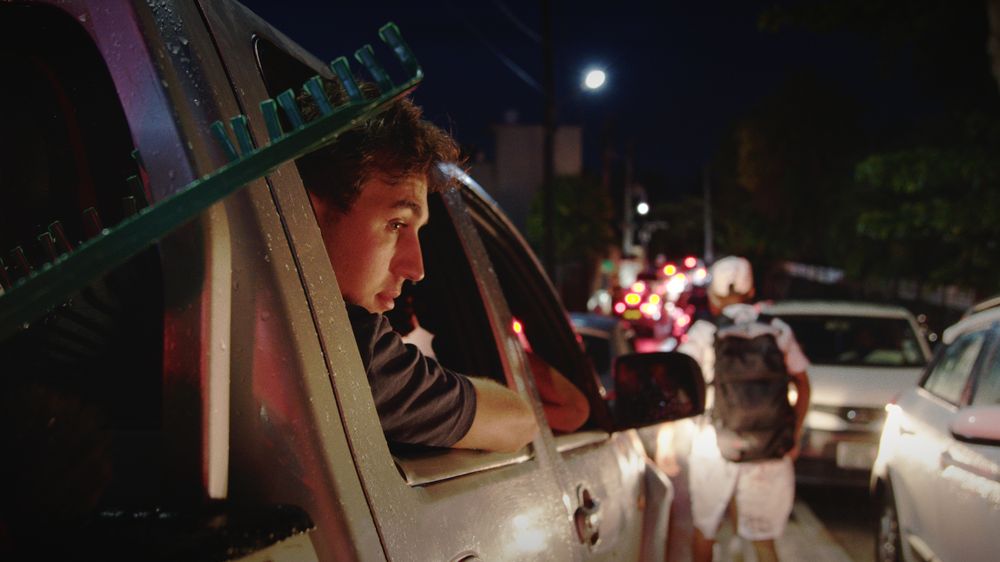 Henri leans out of a pickup window in evening traffic, street lights and brake lights behind