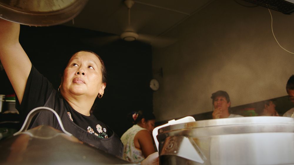A woman lifts the lid of a large pot in a busy kitchen