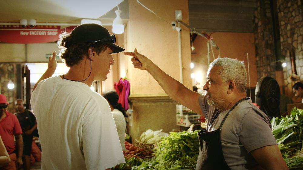 Henri talks with a stall holder who points at him, both smiling, at Port Louis market