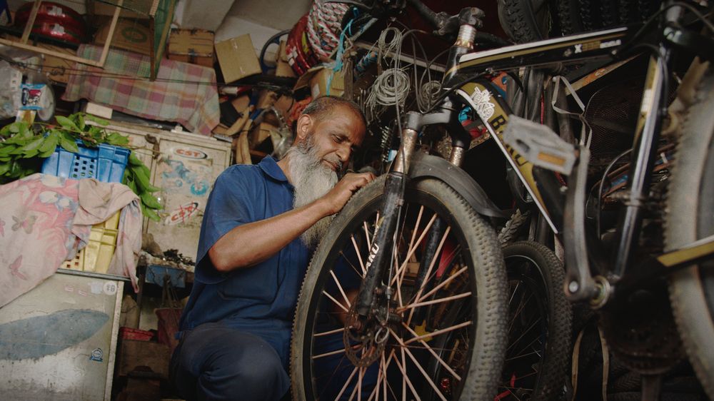 A bike mechanic with a long white beard works on a wheel in a workshop crowded with bikes