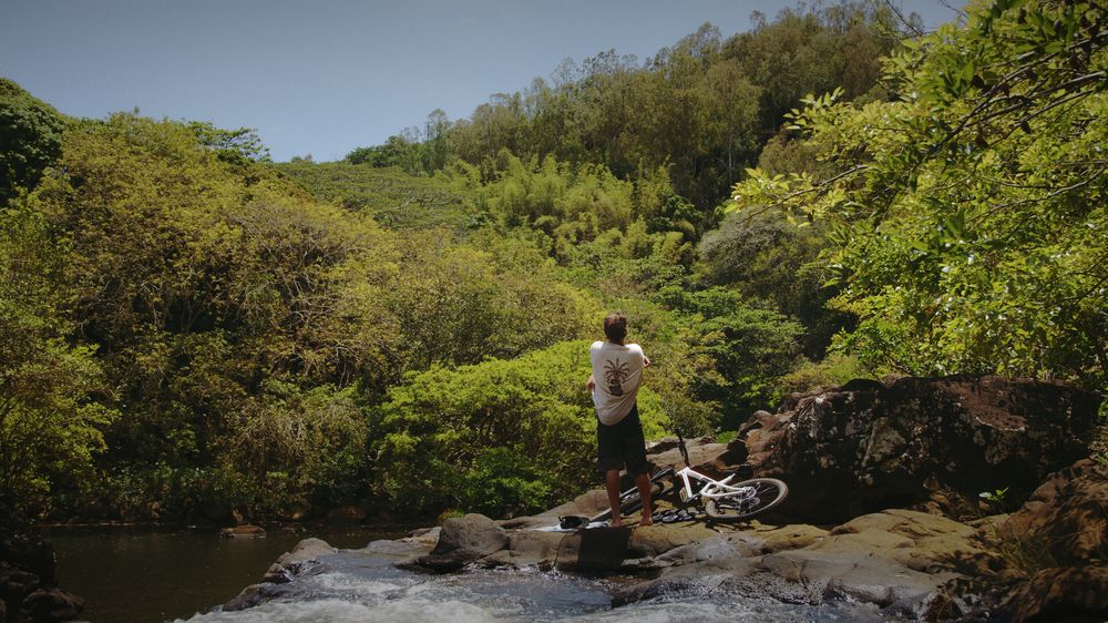 A rider rests his bike on the rocks beside a jungle river