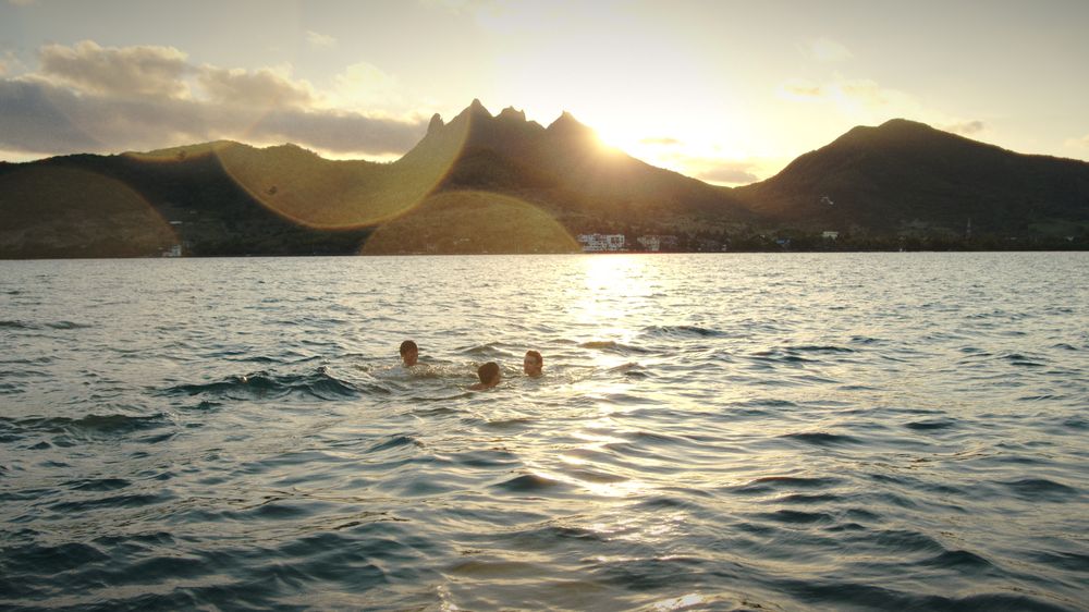 Three friends swim in the sea at sunset with jagged mountains behind