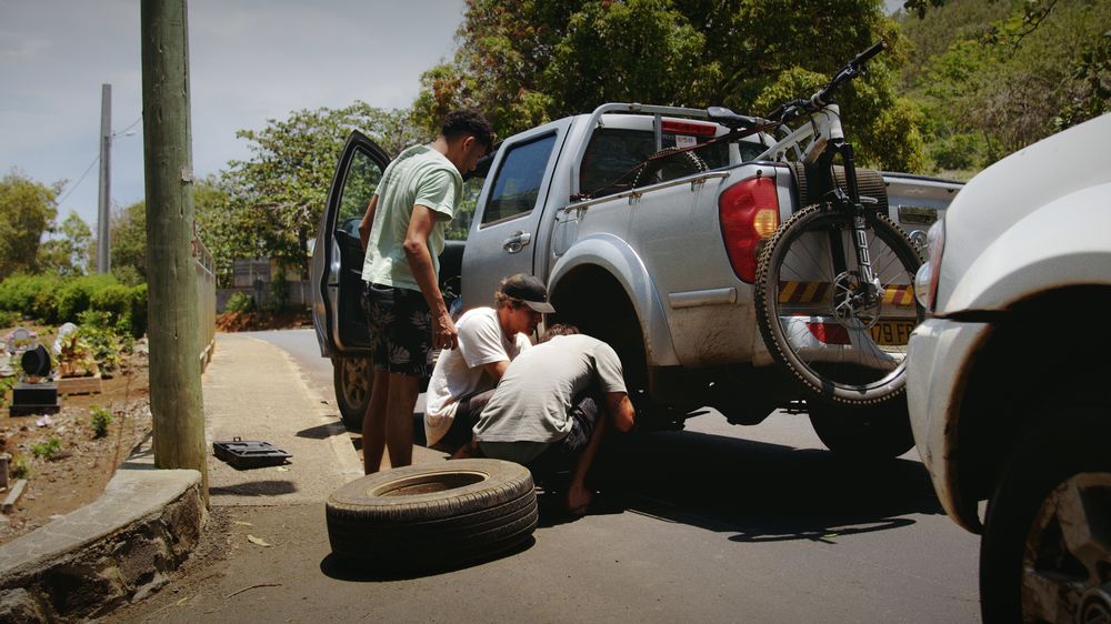 Friends change a wheel on the roadside next to a pickup with a bike on the back
