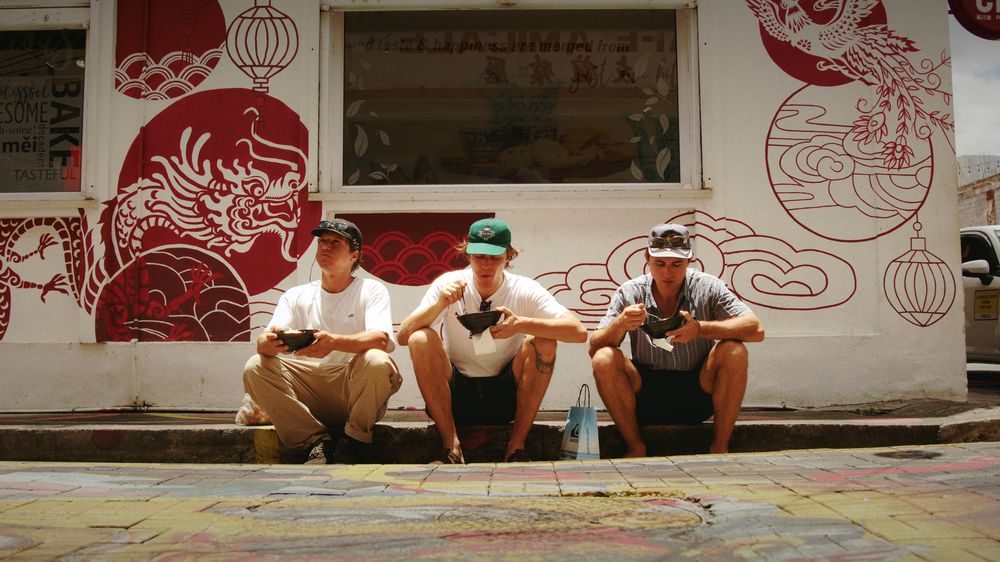 Three friends sit on a step in front of a white wall painted with red dragons and lanterns