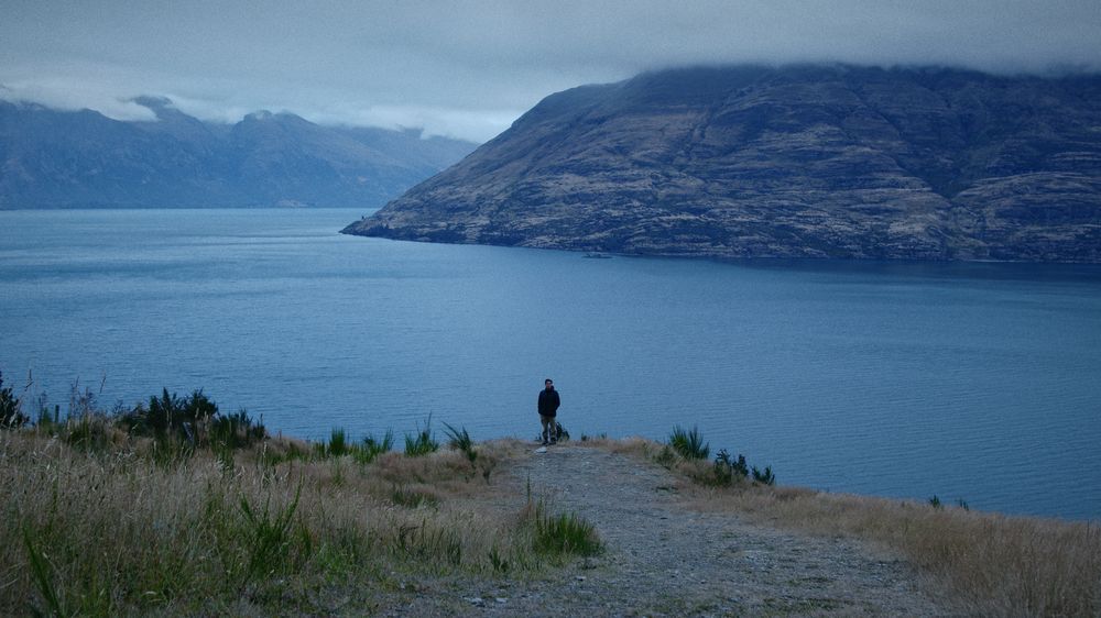 A lone figure with a bike stands on a point above a deep blue lake, mountains under low cloud
