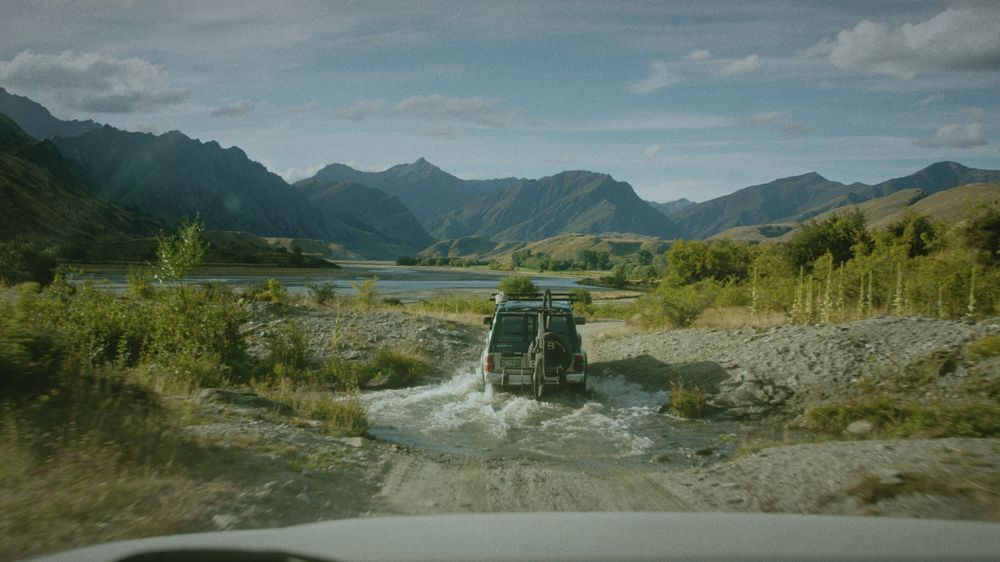An old Land Rover fords a braided river in a wide mountain valley