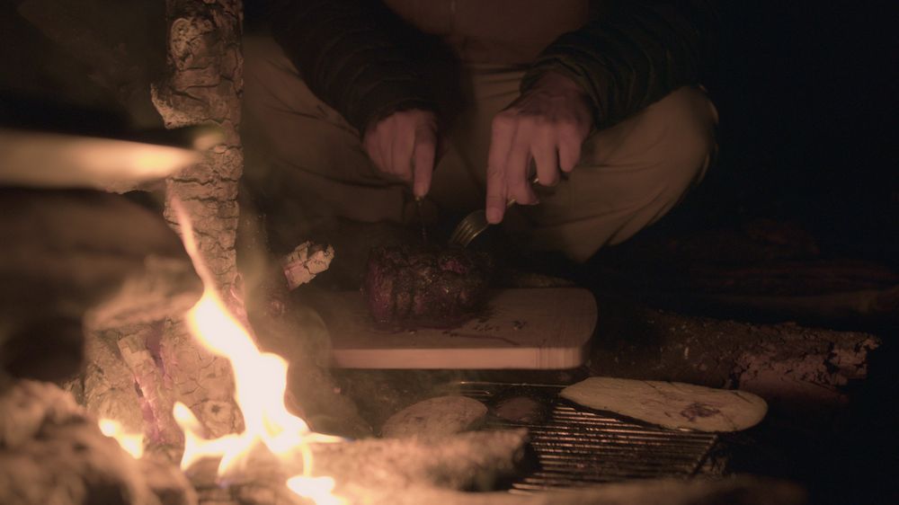 Hands cutting meat beside an open fire at night