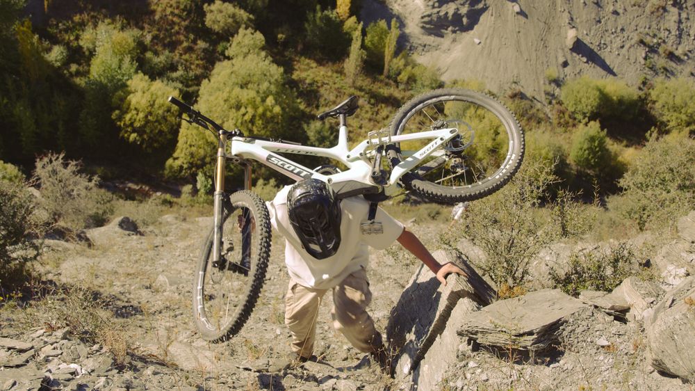 Henri carries his bike up a scree slope, autumn trees behind